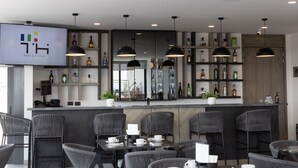Een moderne bar met hoge krukken, een counter waarop een tv staat met de naam van het hotel, en flessenrekken vol met drank.