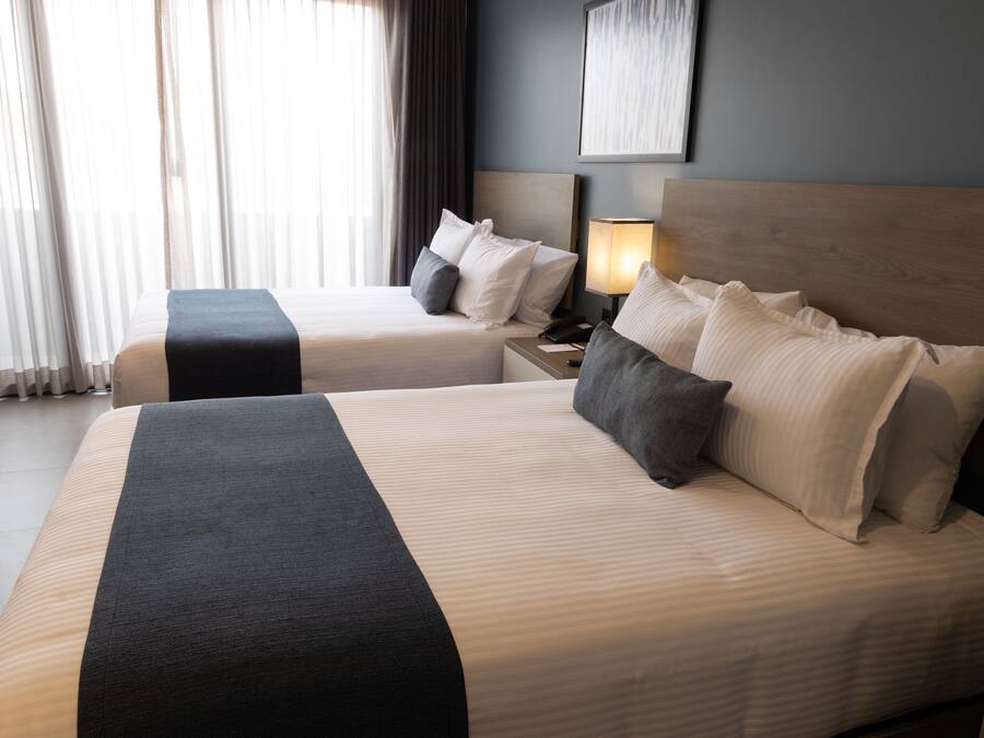 A hotel room with two beds, a nightstand with a lamp, and a large window with curtains.
