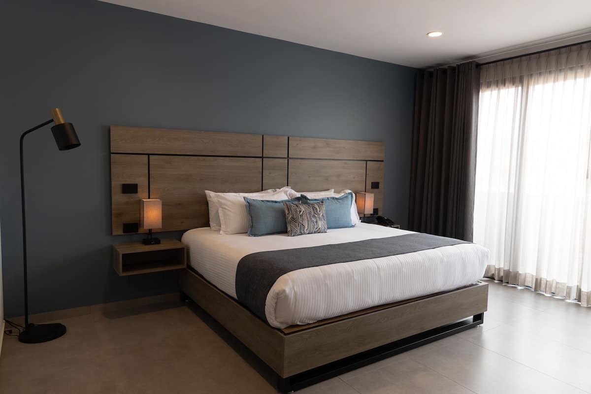 A modern bedroom with a large bed, wooden headboard, bedside lamps, and a view of the outdoors through sheer curtains.