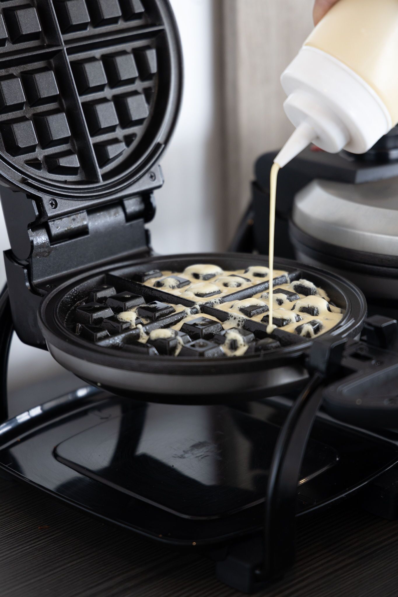 A waffle iron with a grid pattern, and a bottle of batter being poured into it.