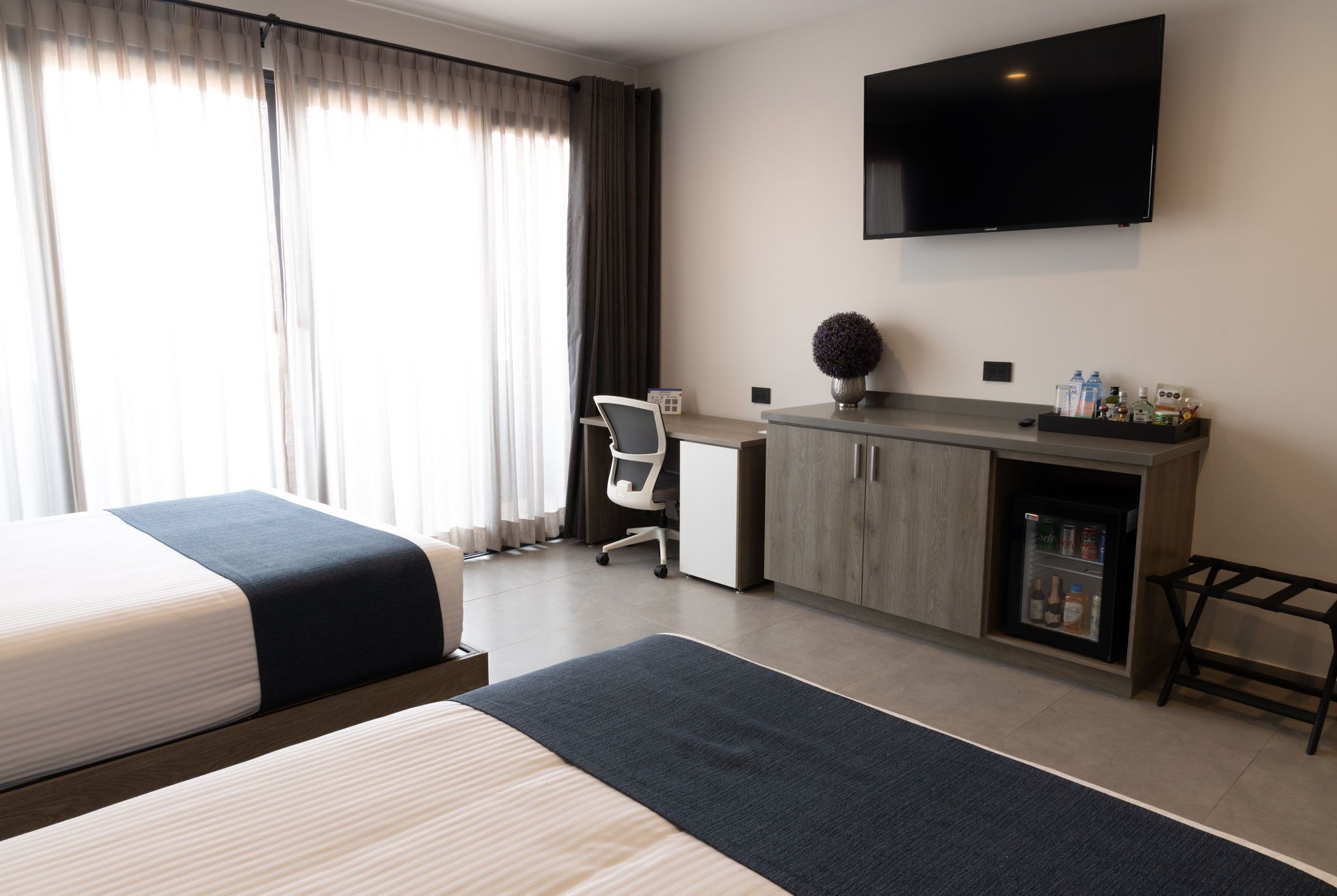 Ein Hotelzimmer mit Bett, Schreibtisch, Stuhl, Fernseher, Minibar und einem Fenster mit Vorhängen.