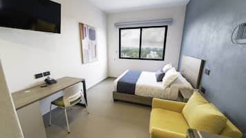 Una habitación de hotel con una cama, un escritorio, una silla, un sofá amarillo, un televisor montado en la pared y un ventanal con vista a la ciudad.