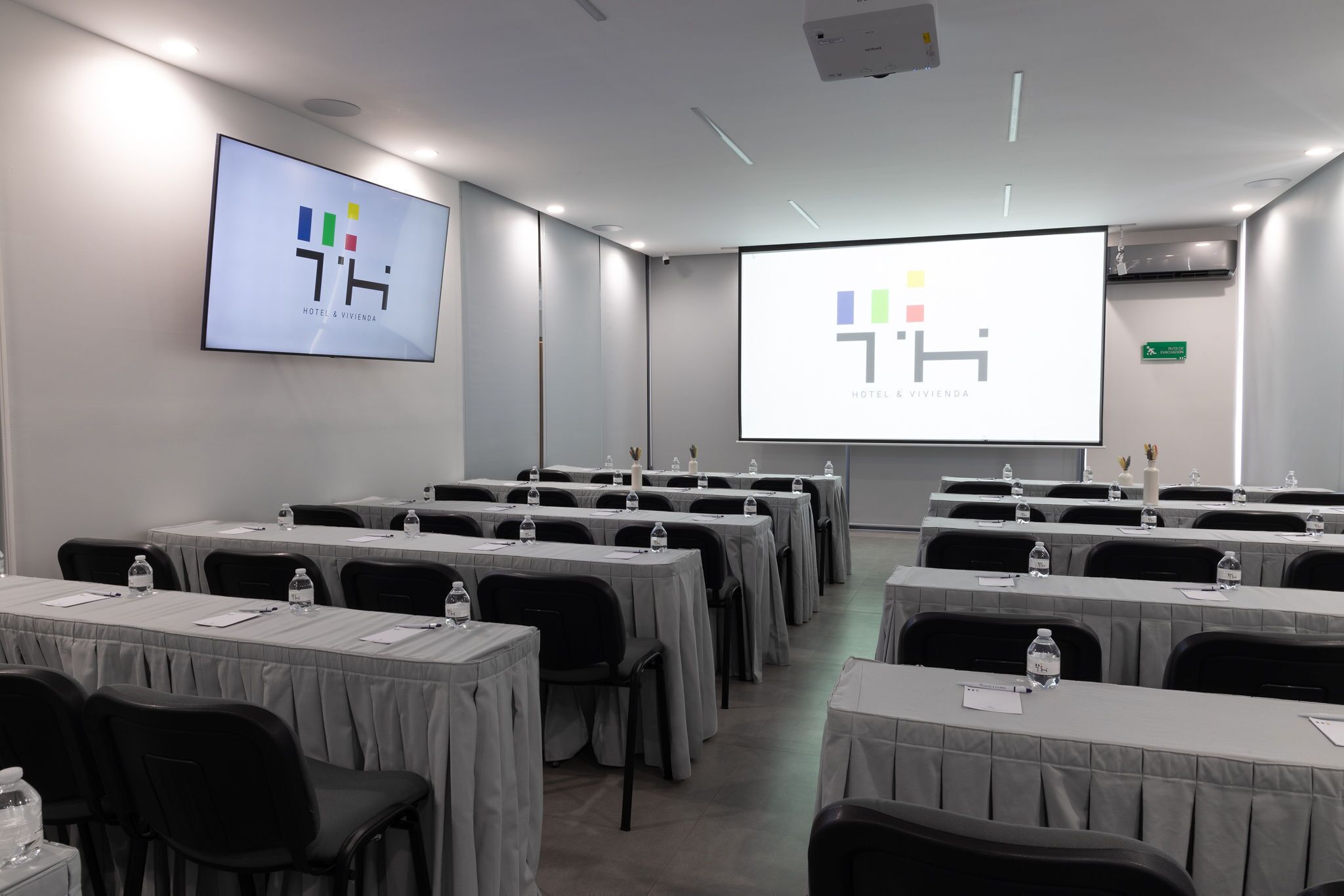 Un salón de conferencias con mesas, sillas y una pantalla proyectora mostrando el logotipo de un hotel.