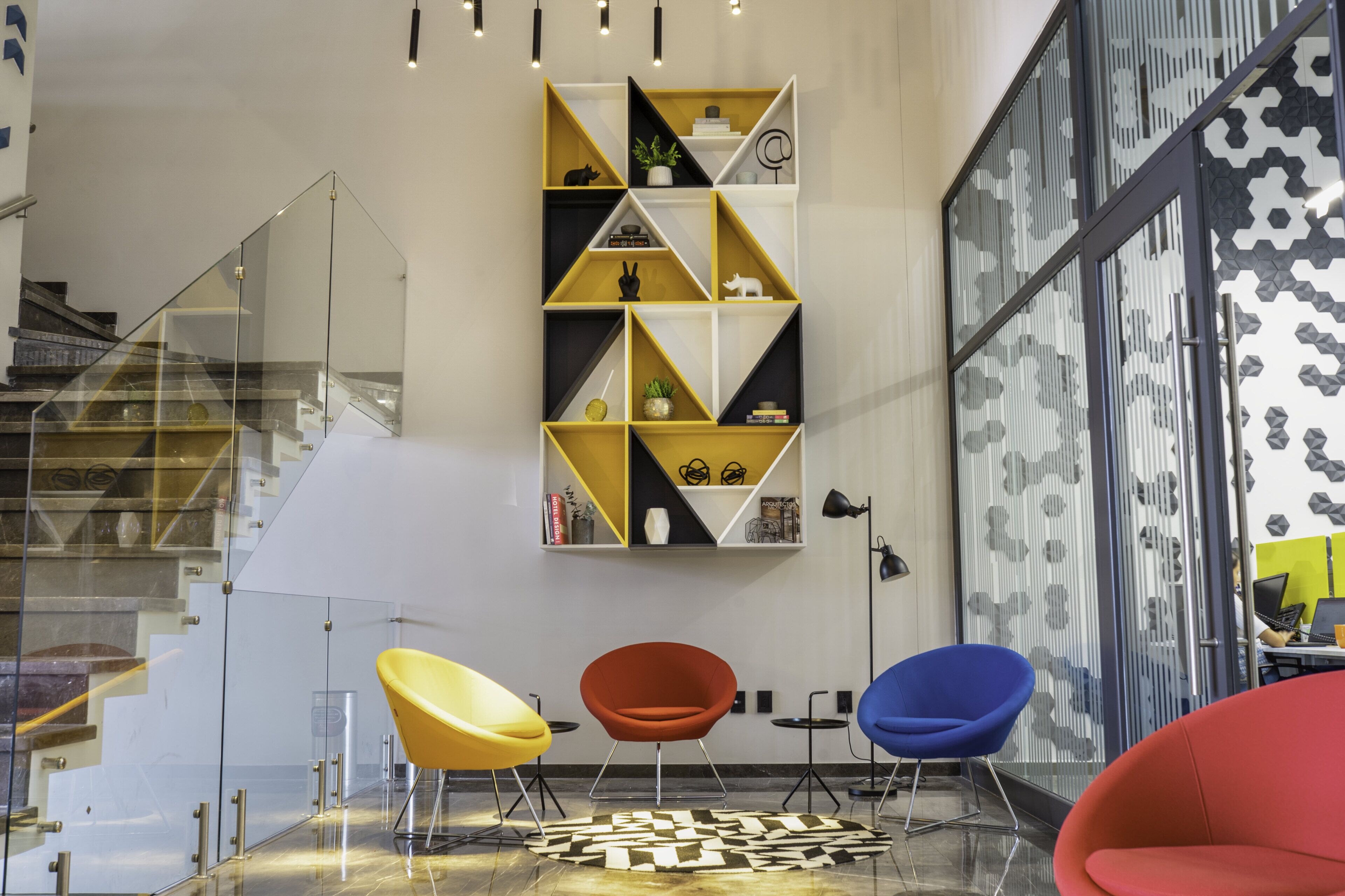 Un interior moderno con un alfombra de diseño geométrico, una escalera con barandillas de vidrio y un mueble de pared con varios objetos decorativos.
