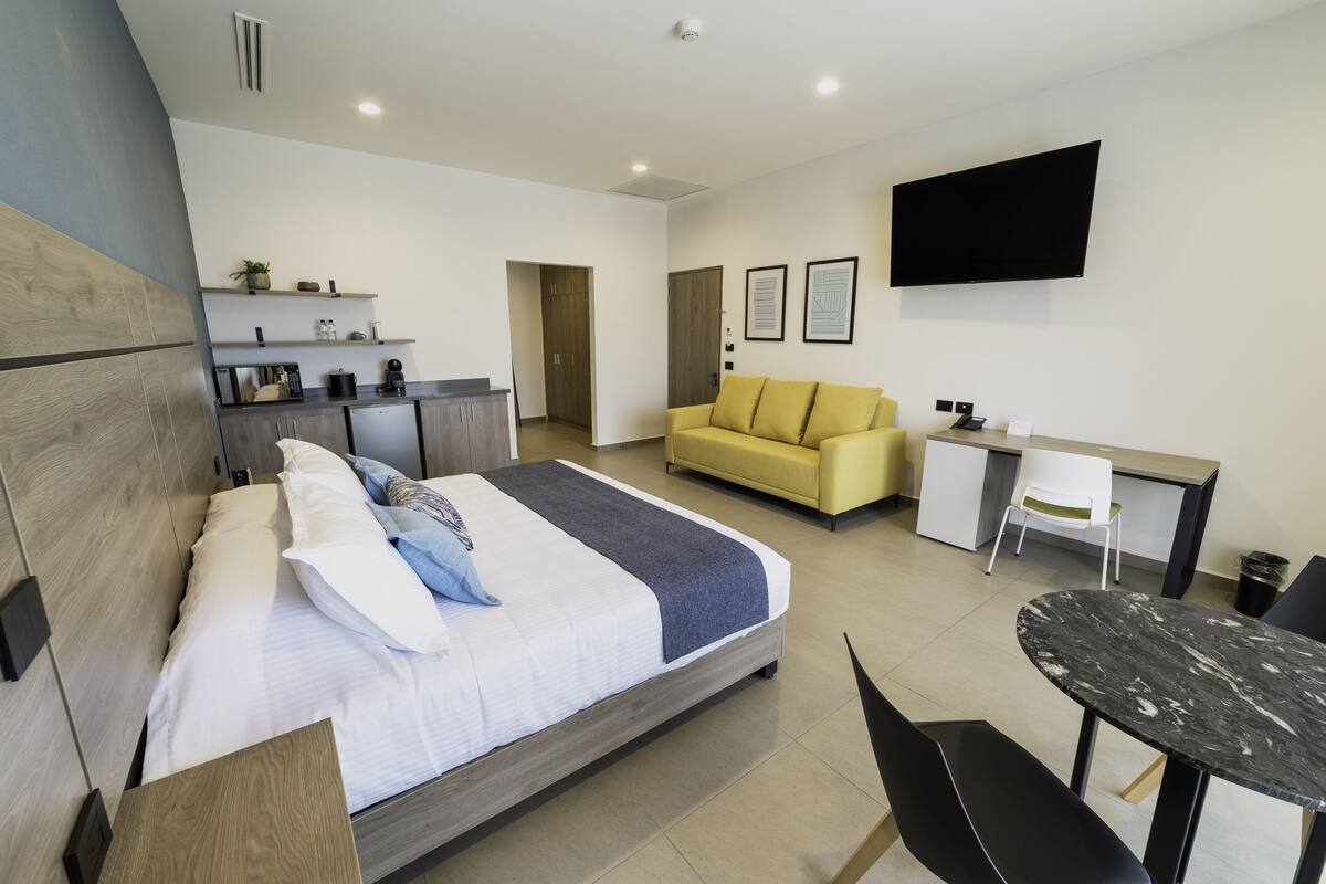 A modern hotel room with a bed, a sofa, a desk, a TV, and a dining table.