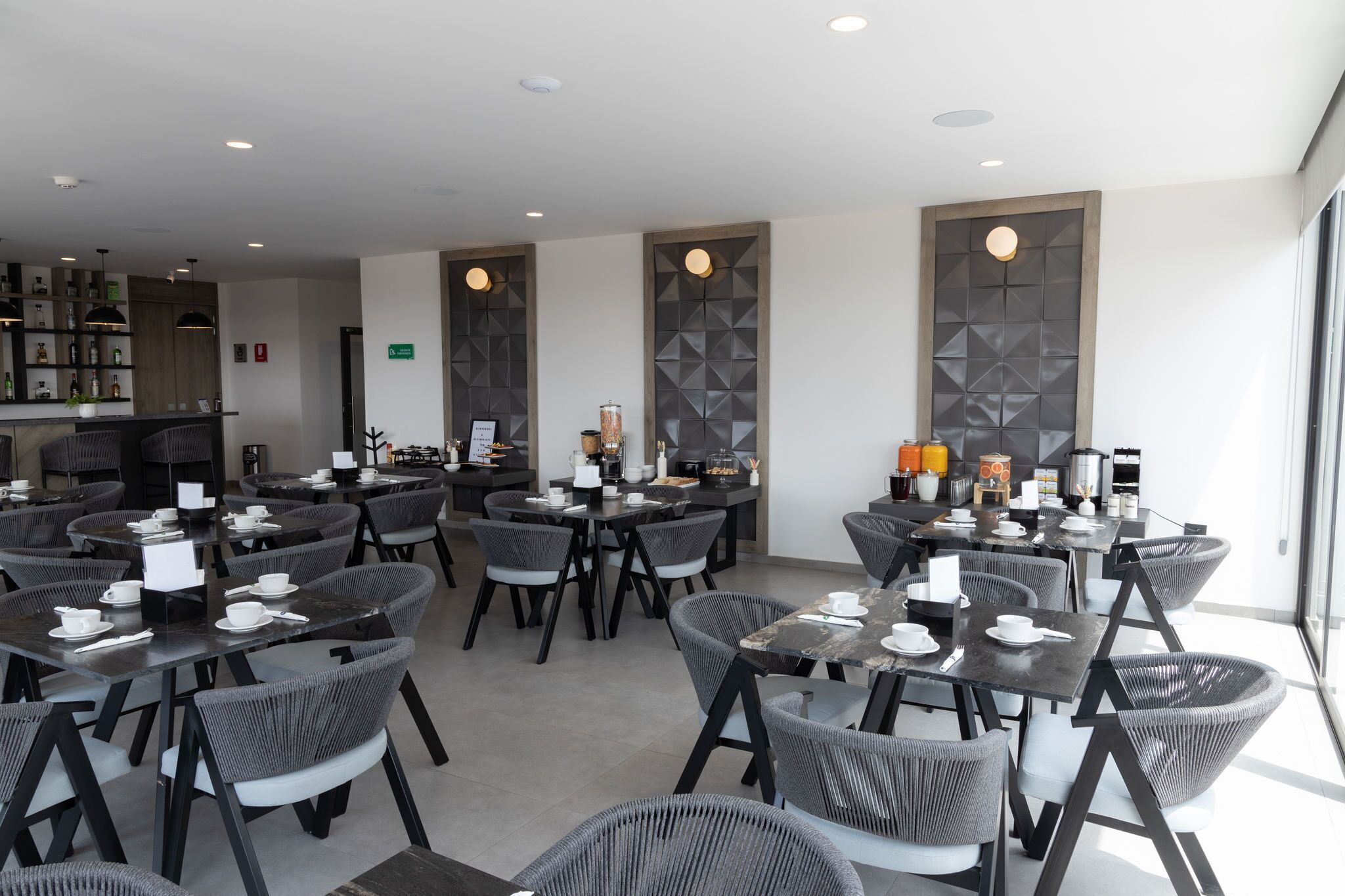 A modern restaurant with round tables and grey chairs, a bar area with shelves of bottles, and a large window providing natural light.