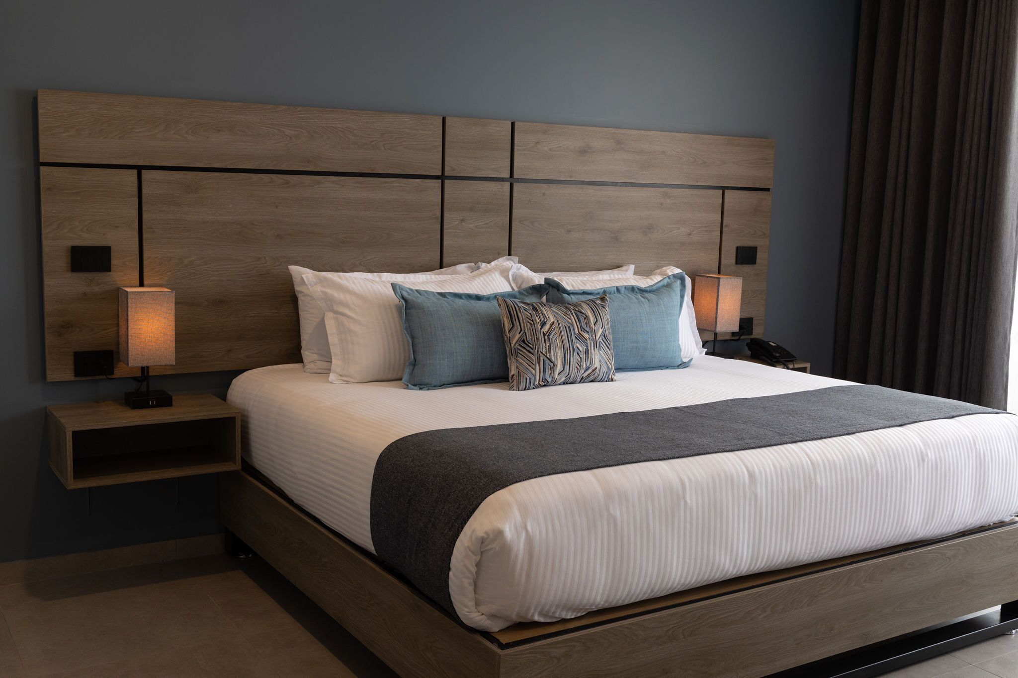 Una cama bien hecha con cabecera de madera, dos lámparas de noche y una mesita auxiliar.