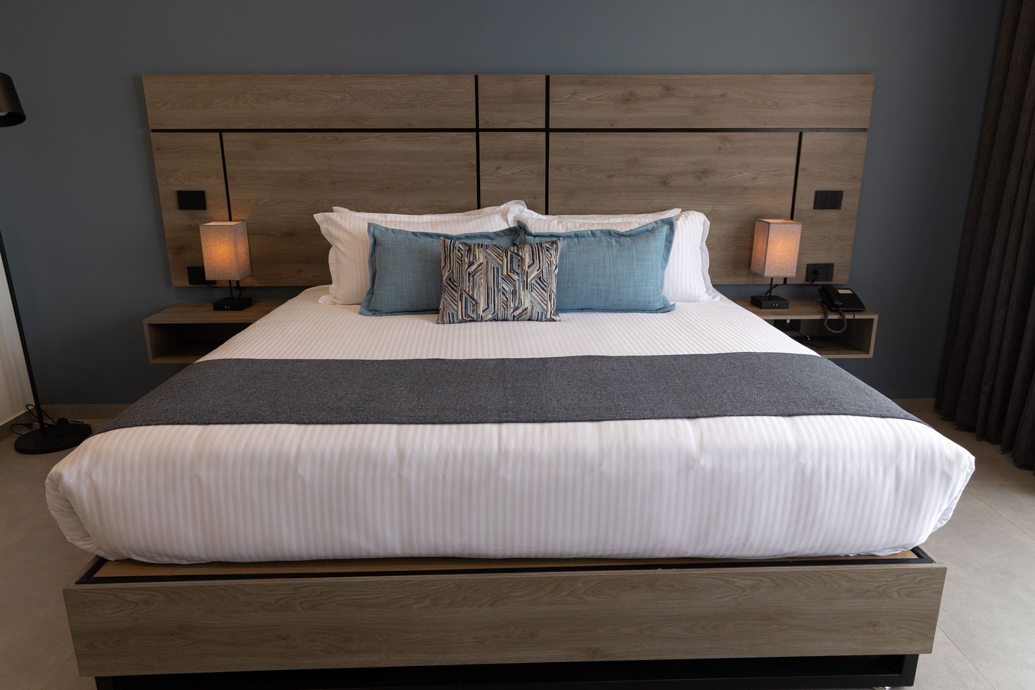 A modern hotel room with a large bed, two bedside tables, and a headboard with a wooden finish.