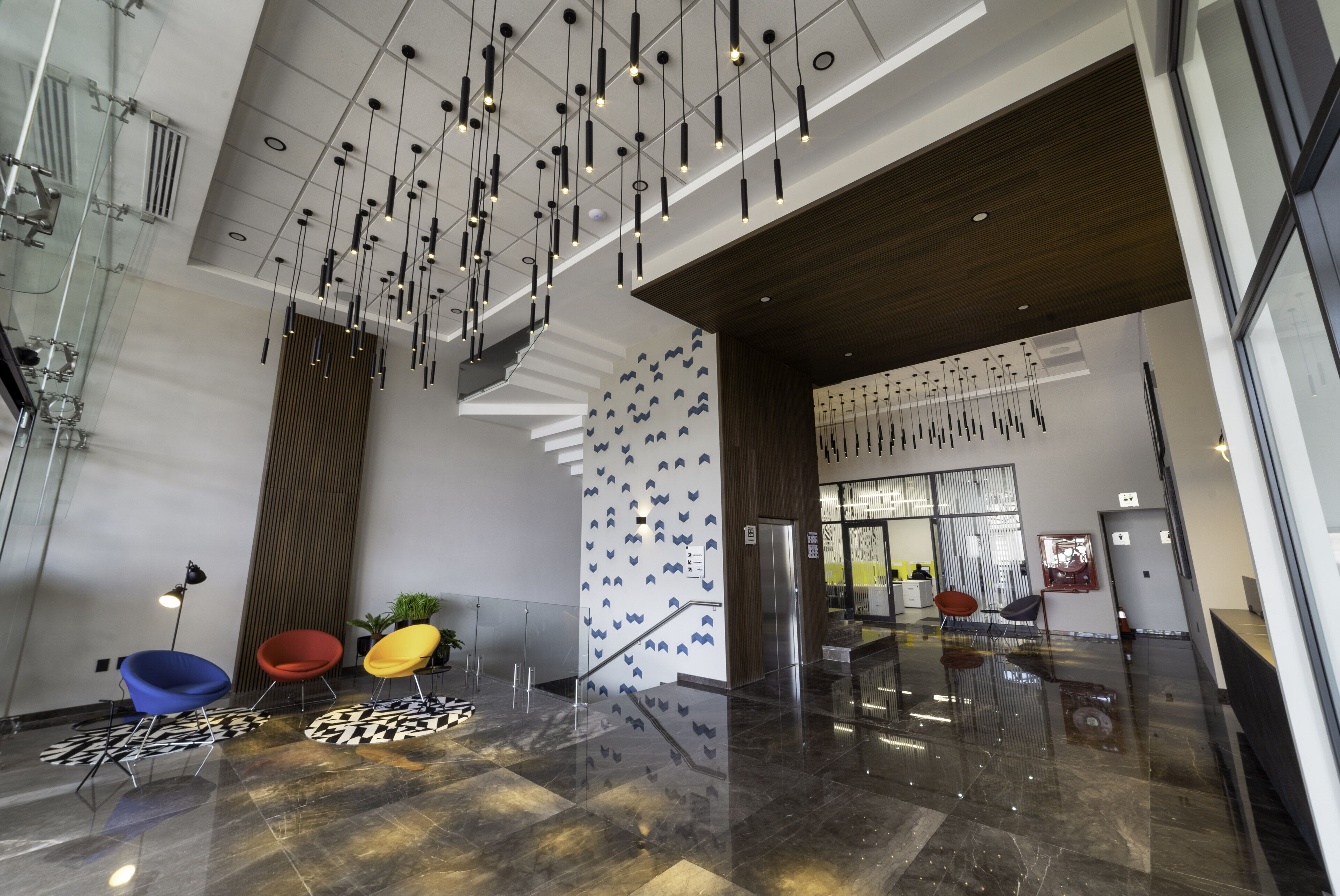 A spacious lobby with a modern design, featuring a high ceiling with hanging lights, a staircase, and a patterned wall.
