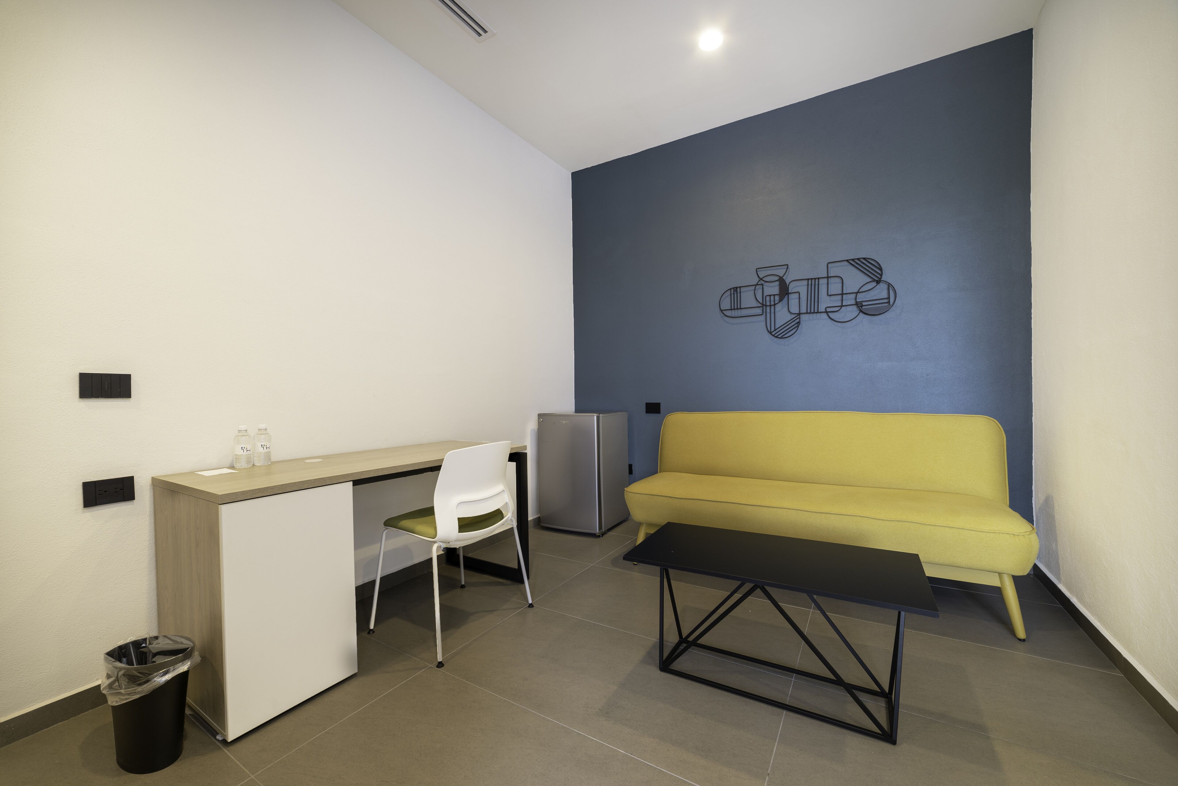 Una habitación compacta con kitchenette, un sofá amarillo, una silla blanca y una mesa negra.
