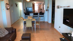 Un saló amb un sofà, otomans, una zona de menjador amb cadires i una taula, una cuina amb armaris i un frigorífic, i una televisió penjada a la paret.