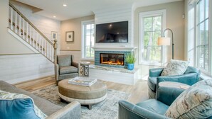 A living room with a fireplace, comfortable seating, a staircase, and large windows. - Traverse City Lakemore Resort, Side by Side Luxury Lakefront Homes on Arbutus Lk (Traverse City)