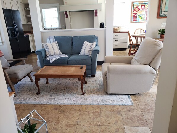 A living room with a sofa, armchair, coffee table, and a kitchen in the background. - Coastal Farm Cottage (Tiverton)