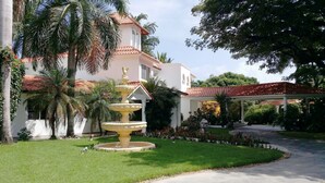 Un jardín bien cuidado con una fuente, palmeras y un paseo cubierto que lleva a un edificio con tejado de baldosas.