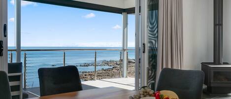A room with a wooden table, a grill with food, a view of the sea, and a chimney.