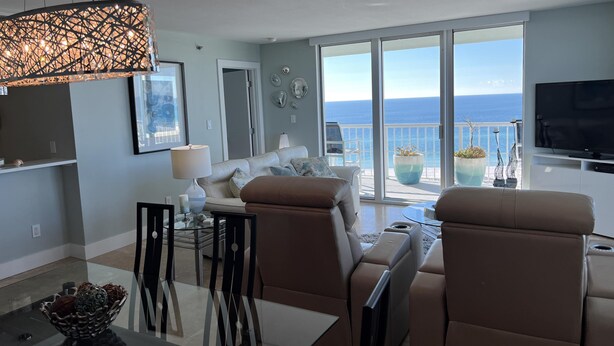 A modern living room with a sofa, chairs, a dining table, and a TV. There is a balcony with a view of the sea. - Luxurious 15th Floor Penthouse, Water Views -15B (Pensacola Beach)