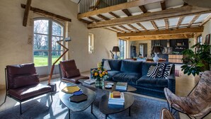 A spacious living room with a dark blue sofa, leather armchairs, and a coffee table with books and a vase of flowers. A wooden ceiling and a view of the kitchen are visible.