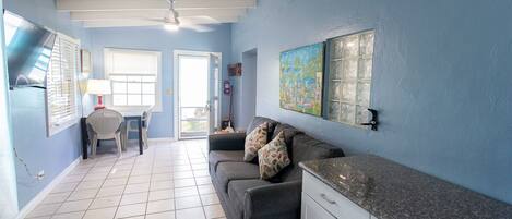 Quarto com parede azul, sofá cinza, mesa de jantar com cadeiras e área de cozinha com armários.