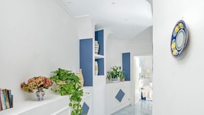 A hallway with white walls, blue accents, and a blue-tiled floor. There are potted plants and a vase with flowers on a shelf. - Villa Blumarine - Private villa with pool and sea views (Positano)