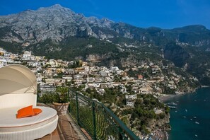 Una cittadina costiera con vista sulle montagne, un balcone con una sedia bianca e un cuscino arancione, e una spiaggia con delle barche.