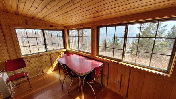 A cabin interior with a wooden ceiling, a red table, and four chairs. Large windows provide views of trees and a forested area. - Superior Reflections Cabin 3 (Schroeder)