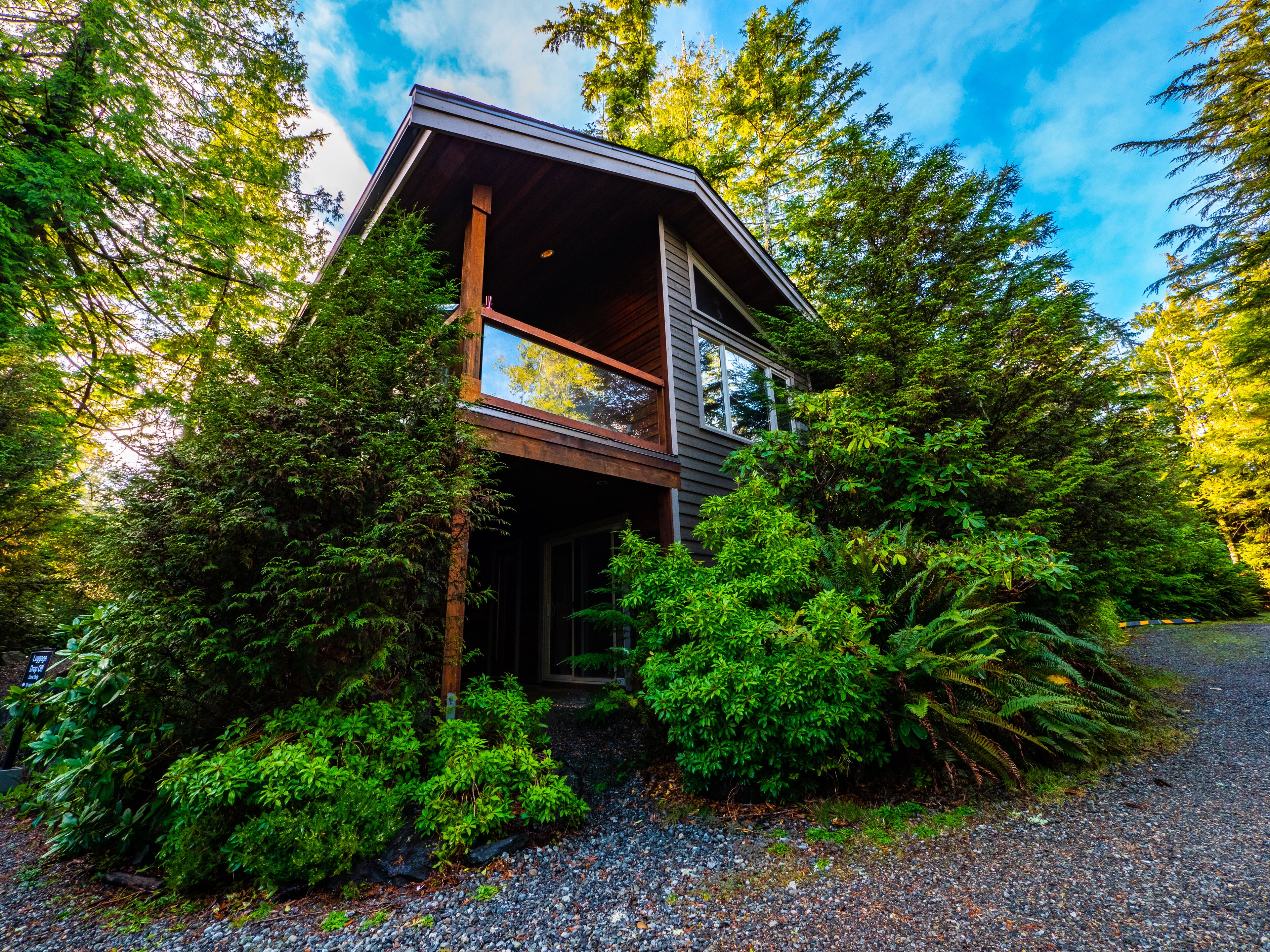 A two-story cabin surrounded by dense forest, with a gravel driveway leading to the entrance.