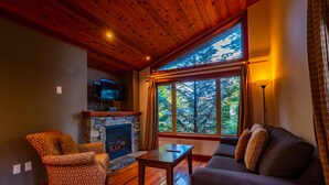 A cozy living room with a stone fireplace, wooden flooring, a television mounted above it, a sofa, and a large window offering a view of trees.
