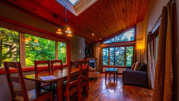 A spacious room with a wooden floor, a dining table with chairs, a sofa, and large windows offering a view of trees.
