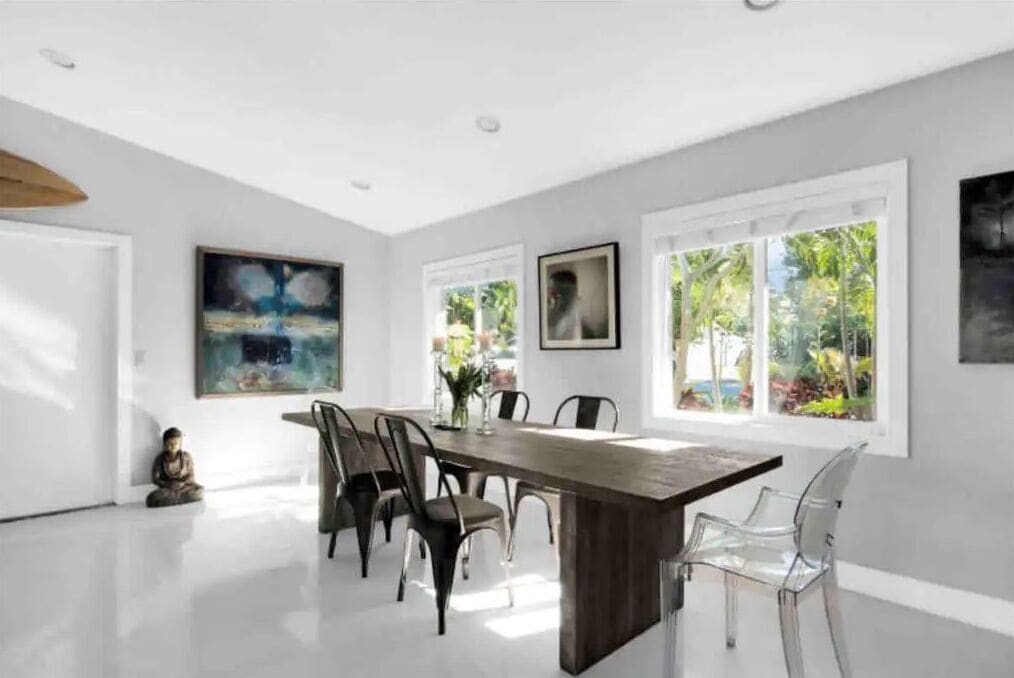 A modern dining room with a long wooden table, clear chairs, and large windows offering a view of greenery.