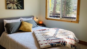 Un dormitorio con cama, almohadas, una manta, una mesita de noche con lámpara, un cuadro en la pared y una ventana con vistas a árboles.