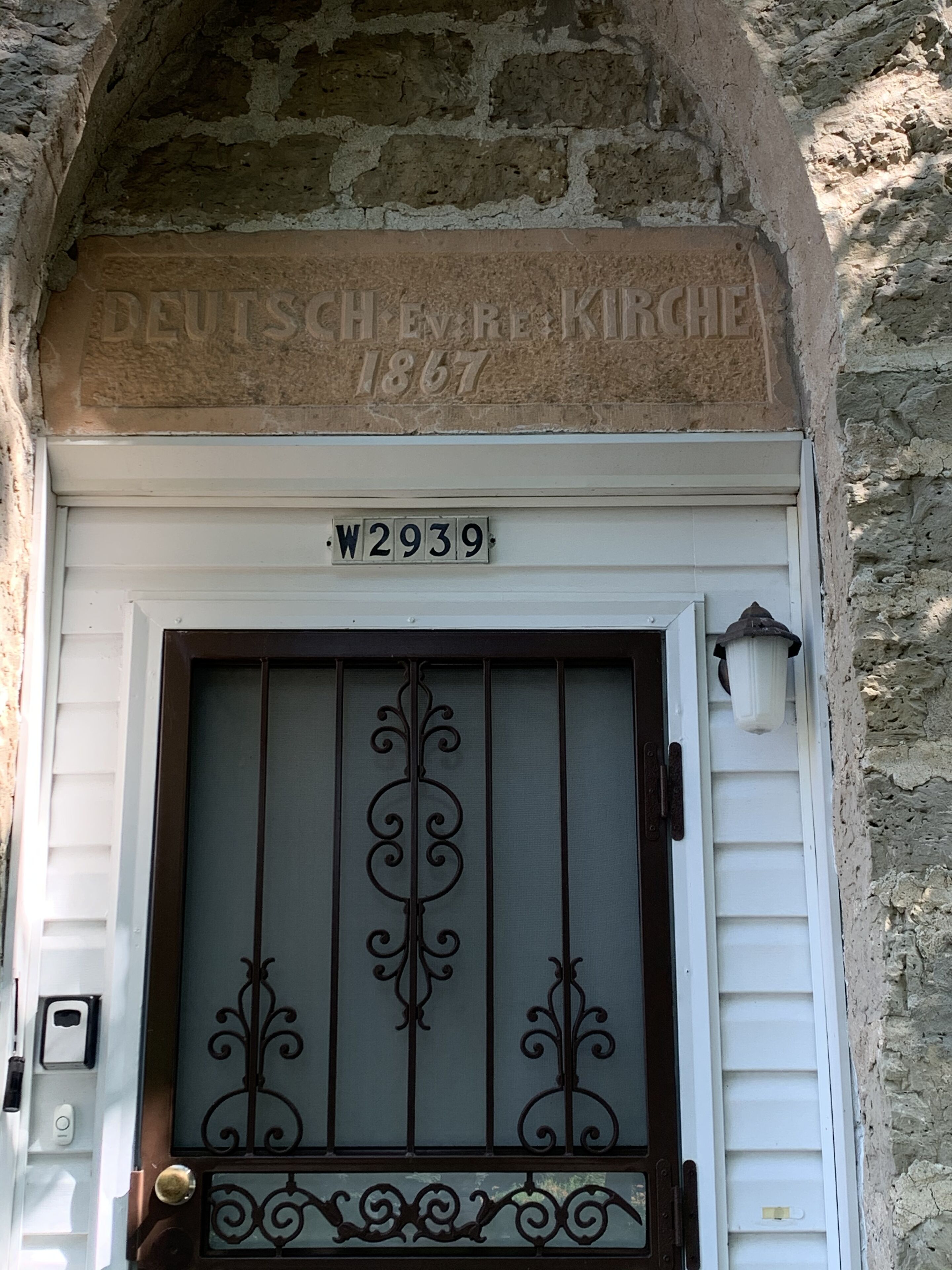 L’entrée d’un immeuble est ornée d’une arche en pierre, d’une enseigne indiquant « DEUTSCHER BAKKERIE 1867 » et d’une porte surmontée d’un portail en métal.
