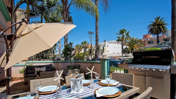An outdoor patio with a dining table set for four, surrounded by wicker chairs, a grill, and palm trees. - Lucy's Beach House, (Oceanside)