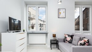 Un salon moderne avec un canapé gris, un téléviseur à écran plat posé sur un meuble blanc, une petite table d’appoint avec une lampe, et une fenêtre donnant sur des immeubles.