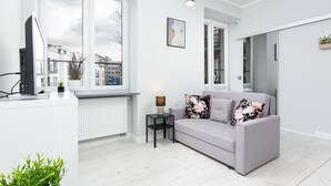 Un salon moderne avec un canapé gris, une petite table d’appoint noire sur laquelle se trouve une lampe, et un téléviseur posé sur un meuble blanc. Une grande fenêtre offre une vue sur les bâtiments environnants.