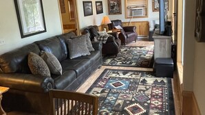 A living room with a dark leather couch, a wooden dining table with paw print coasters, a patterned rug, and a ceiling fan. - Northwoods waterfront Rhinelander Cabin getaway (Rhinelander)