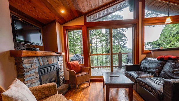 Ein gemütliches Wohnzimmer mit Kamin, Holzböden, einem Ledersofa und großen Fenstern mit Blick auf Bäume.