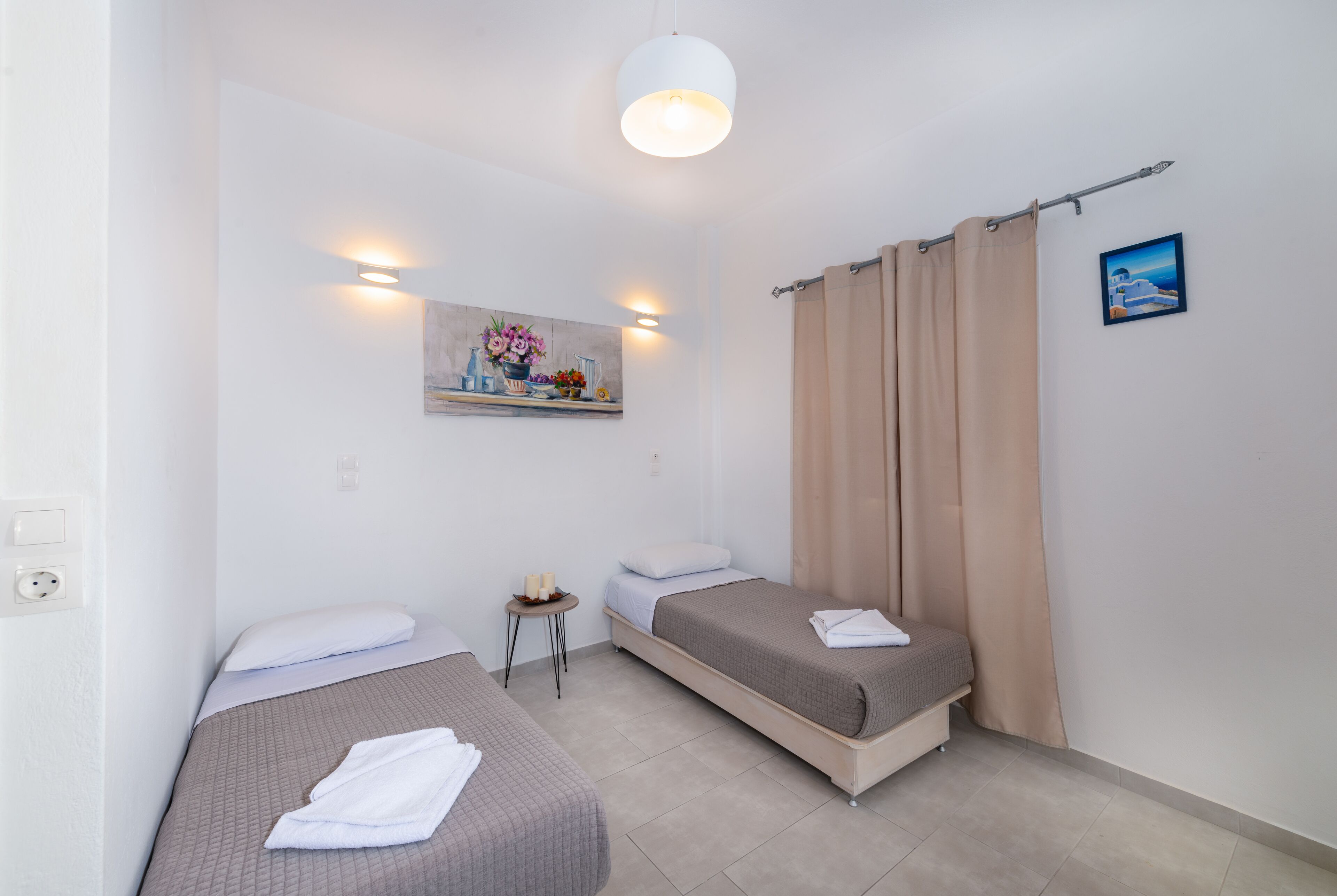 Habitación de hotel con dos camas individuales, una mesita con un florero y un cuadro en la pared, además de una cortina en la ventana.