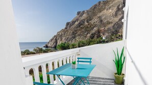 A balcony with a table and chairs, a potted plant, and a view of the sea and rocky cliffs. - Aislin House: Where Beachfront Bliss Meets Santorini Charm! (Santorini)