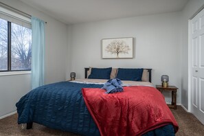 Un dormitorio con cama, ventana con cortinas, mesita de noche con lámpara y un cuadro en la pared.