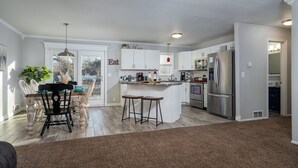 A modern kitchen with white cabinets, a central island with stools, stainless steel appliances, and a dining area with a table and chairs.