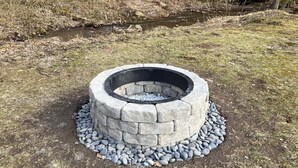 Uma lareira de pedra com design circular em pedra e um aro de metal, cercada por uma borda de pedra e uma camada de pedras planas.
