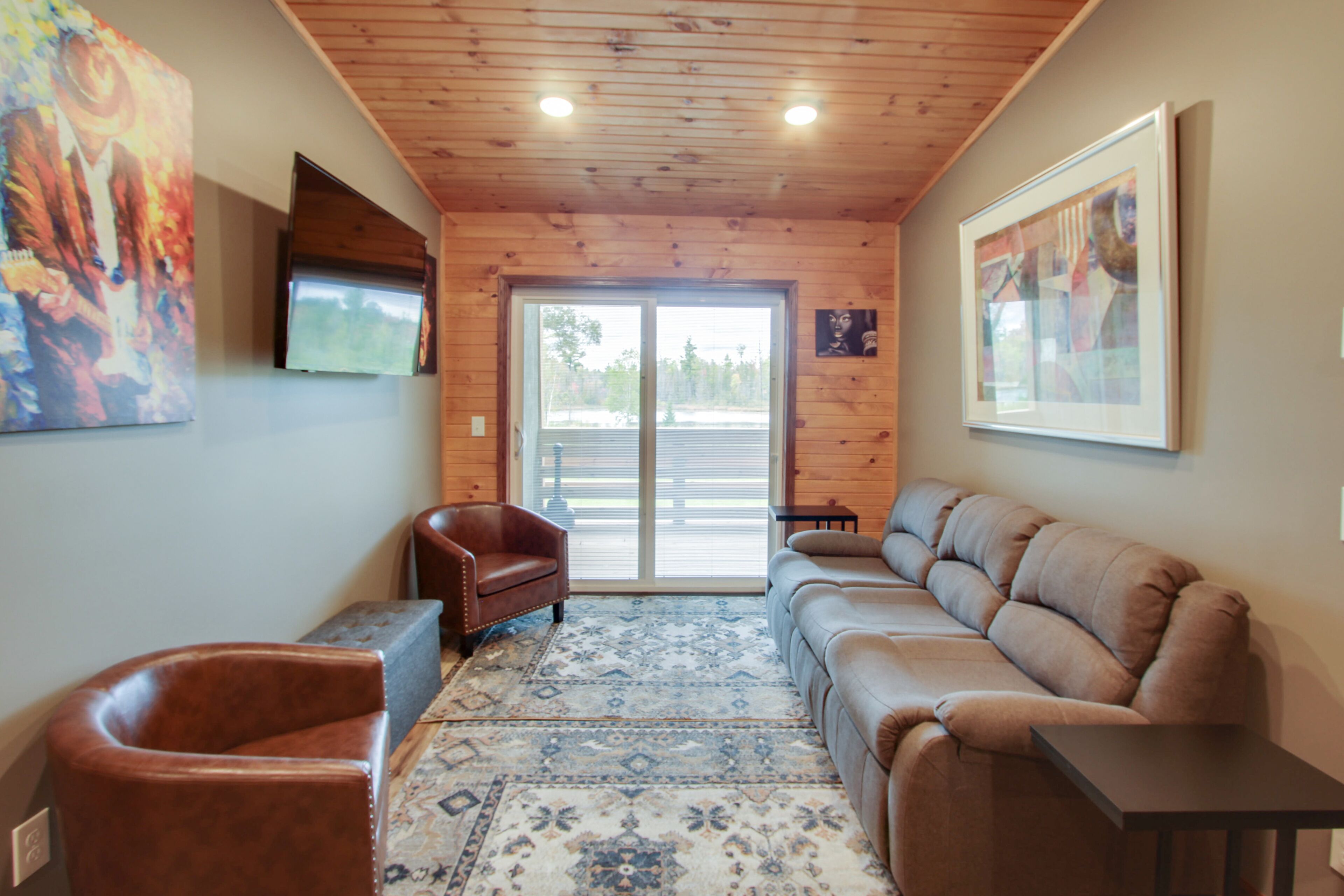 Una habitación con techo de madera, un ventanal grande, un sillón de cuero marrón, un sofá color beige y un tapete estampado.