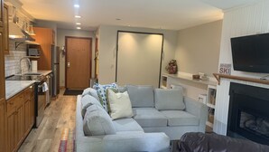 A modern living room with a sofa, a fireplace, and a kitchen area with wooden cabinets and a built-in oven. - Sugarloaf Ski-In/Ski-Out Condo – Golf, Bike & Hike (Carrabassett valley)