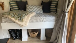 A cozy bed with a patterned bedspread, multiple pillows, and a throw blanket. There are storage baskets under the bed and a pair of boots beside it. - Sugarloaf Ski-In/Ski-Out Condo – Golf, Bike & Hike (Carrabassett valley)