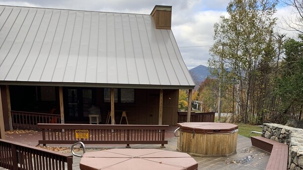 A wooden deck with hot tubs, a stone wall, and a building with a metal roof. - Sugarloaf Ski-In/Ski-Out Condo – Golf, Bike & Hike (Carrabassett valley)