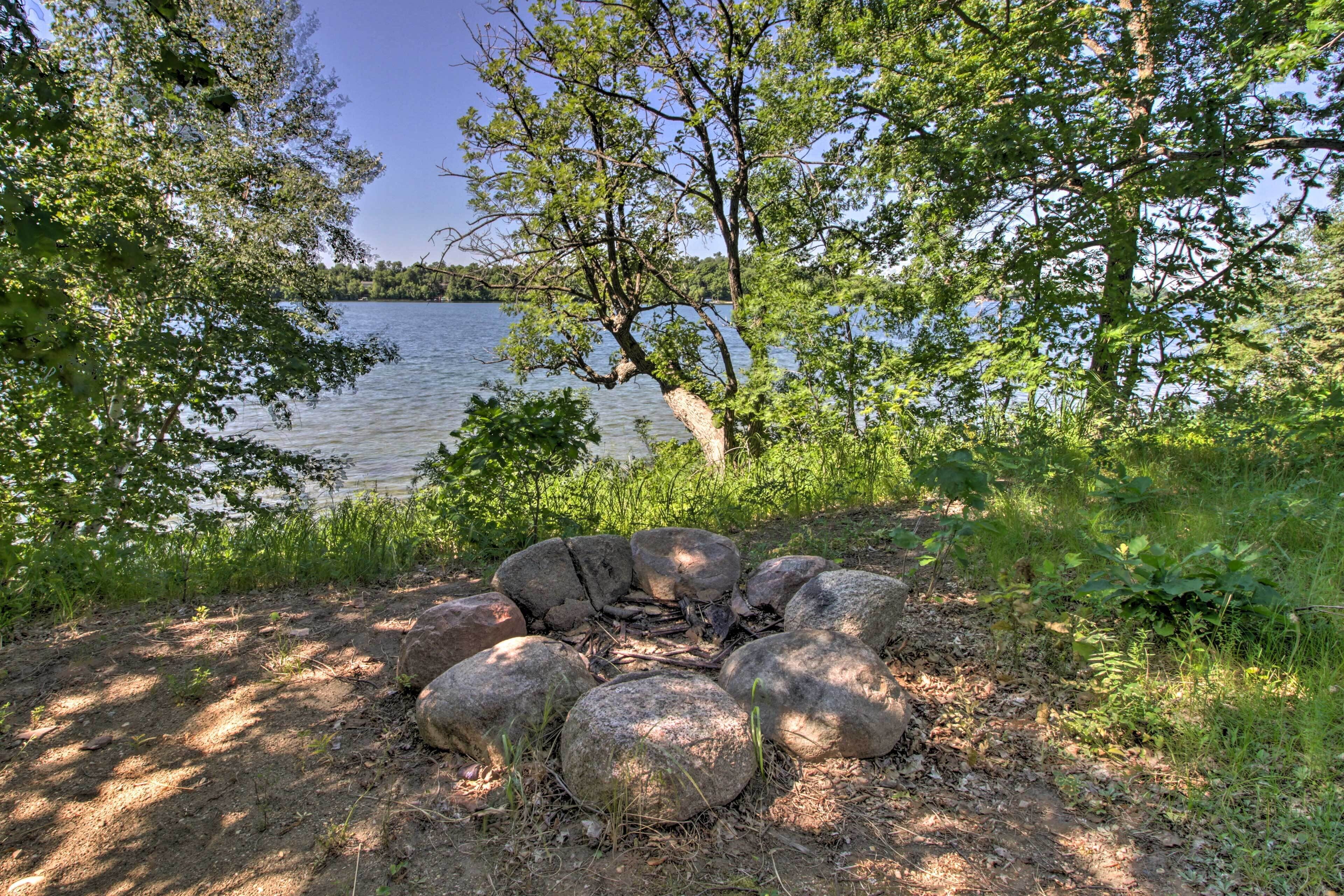 A natural outdoor setting with a fire pit surrounded by rocks, a lake in the background, and trees.