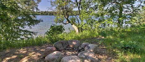 A natural outdoor setting with a fire pit surrounded by rocks, a lake in the background, and trees.