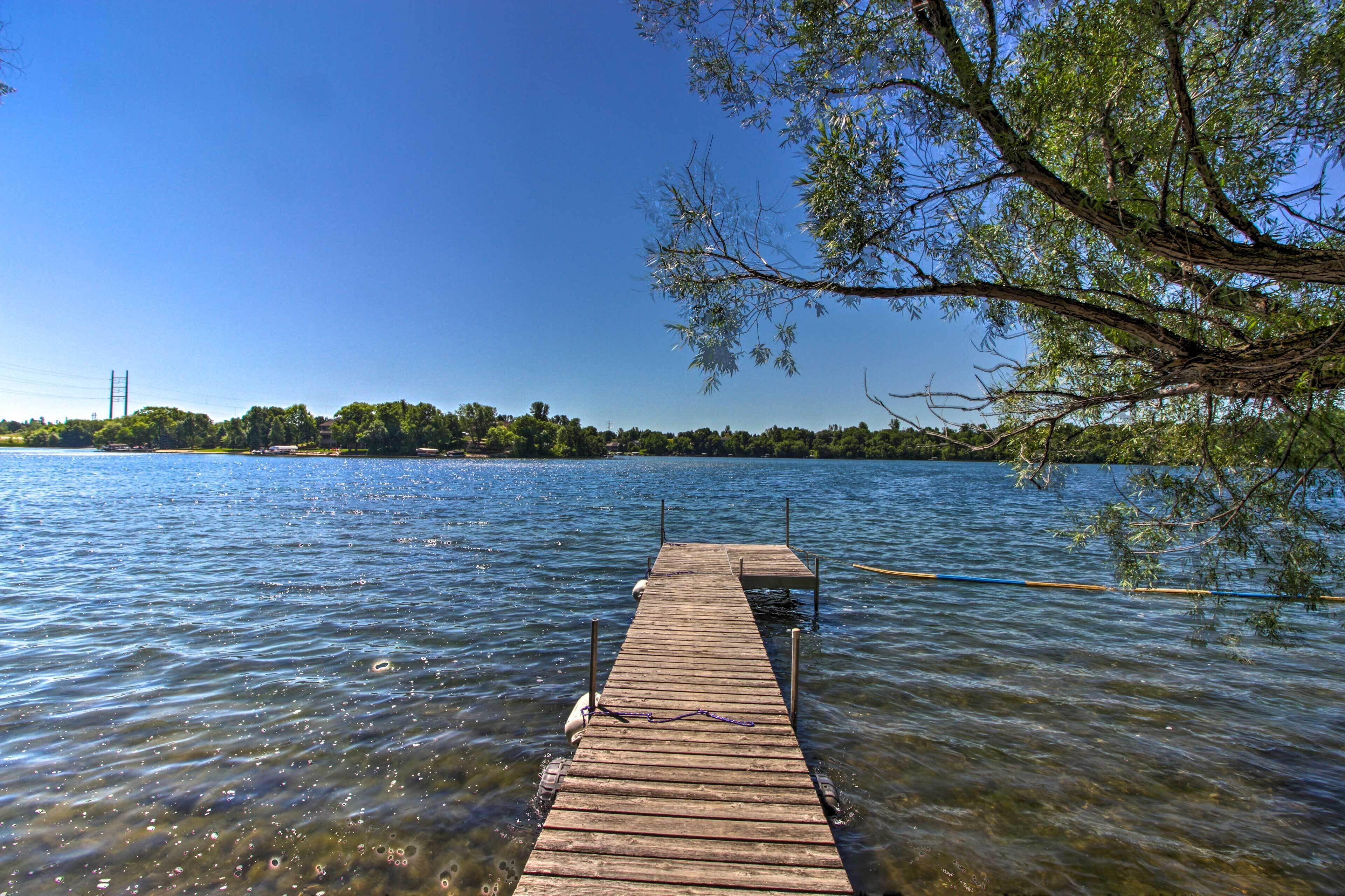 A wooden pier extending into a calm lake with clear water, surrounded by trees and a distant shoreline.