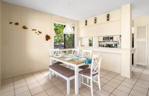 A dining area with a white table set for four, a kitchen with modern appliances, and a large window with a view of greenery.
