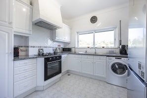 A modern kitchen with white cabinetry, a built-in oven, a microwave, a washing machine, and a window with a view. - CoolHouses Algarve Luz | Blue Ocean View T2 (Luz)